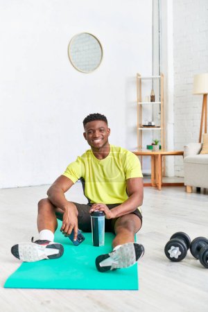 Unbekümmerter afrikanisch-amerikanischer Mann mit Sportflasche und Handy blickt auf Fitnessmatte in die Kamera
