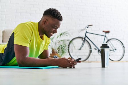 fröhlicher sportlicher afrikanisch-amerikanischer Mann mit Smartphone auf Fitnessmatte neben Sportflasche liegend