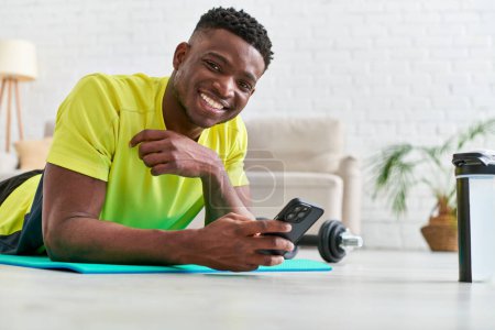 jolly african american man with mobile phone lying down on fitness mat and smiling at camera