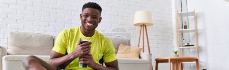 Téléchargez les photos : Heureux sportif homme afro-américain avec bouteille de sport regardant la caméra dans le salon moderne, bannière - en image libre de droit
