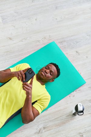young african american man lying on fitness mat and browsing internet on smartphone, top view
