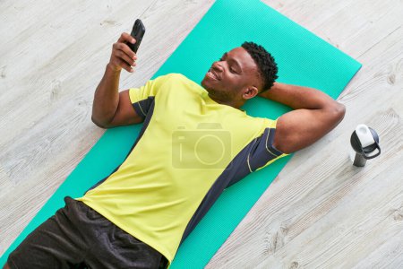 smiling african american man lying on fitness mat and browsing social media on smartphone, top view