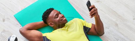 joyful african american guy lying on fitness mat and browsing internet on smartphone, banner