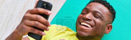 laughing african american man lying on fitness mat and browsing social media on smartphone, banner