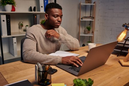 concentrated african american man typing on laptop while working in evening at home, freelancer