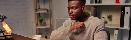 joven afroamericano concentrado freelancer que trabaja en el ordenador portátil en la noche en casa, bandera