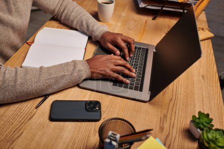 Téléchargez les photos : Vue recadrée de l'homme afro-américain tapant sur ordinateur portable tout en travaillant la nuit à la maison, pigiste - en image libre de droit