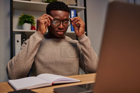 young african american man adjusting eyeglasses near laptop and notebook while working at home