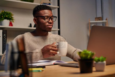 Téléchargez les photos : Focalisé afro-américain pigiste dans des lunettes tenant tasse de café et travaillant sur ordinateur portable à la maison - en image libre de droit