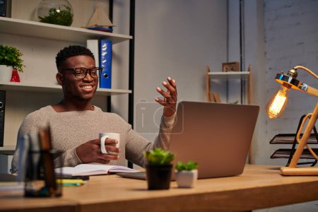 Téléchargez les photos : Heureux homme afro-américain avec tasse de café geste lors d'un appel vidéo au bureau à domicile, pigiste - en image libre de droit