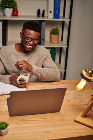 Téléchargez les photos : Homme afro-américain joyeux avec tasse de café rire lors d'un appel vidéo au bureau à domicile, pigiste - en image libre de droit