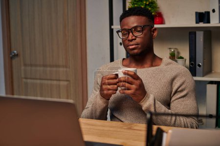 hombre afroamericano reflexivo con taza de café mirando el ordenador portátil mientras trabaja en casa por la noche