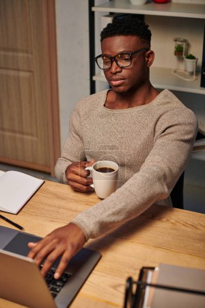 hombre afroamericano concentrado con taza de café trabajando en el ordenador portátil en la oficina en casa por la noche