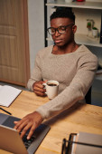 concentrated african american man with coffee cup working at laptop in home office at night tote bag #692608134