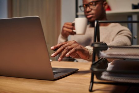 Téléchargez les photos : Jeune pigiste afro-américain avec tasse de café travaillant à l'ordinateur portable dans le bureau à la maison la nuit - en image libre de droit
