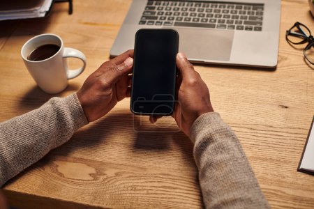vista recortada del freelancer afroamericano que sostiene el teléfono inteligente con la pantalla en blanco cerca del ordenador portátil en casa