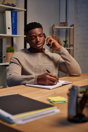 freelancer afroamericano serio hablando en smartphone y escribiendo en notebook, trabajo desde casa