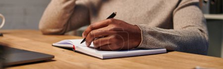 vista recortada del freelancer afroamericano escribiendo en cuaderno mientras trabaja en casa, pancarta