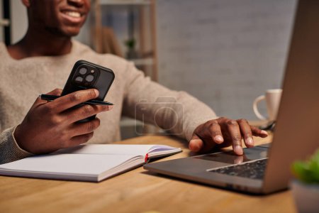 Téléchargez les photos : Heureux homme afro-américain avec smartphone en utilisant un ordinateur portable tout en travaillant à la maison, vue recadrée - en image libre de droit