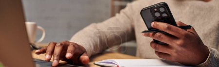 cropped view of african american man with smartphone using laptop while working at home, banner
