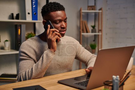 konzentrierter afrikanisch-amerikanischer Freiberufler, der von zu Hause aus arbeitet und in der Nähe seines Laptops mit dem Smartphone spricht