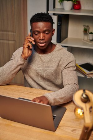 Téléchargez les photos : Jeune indépendant afro-américain concentré travaillant à la maison et parlant sur smartphone près d'un ordinateur portable - en image libre de droit