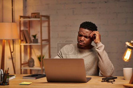 thoughtful african american freelancer looking at laptop in home office at night, problem solving