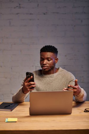 Téléchargez les photos : Freelance afro-américaine grave avec geste smartphone pendant le chat vidéo sur ordinateur portable à la maison - en image libre de droit
