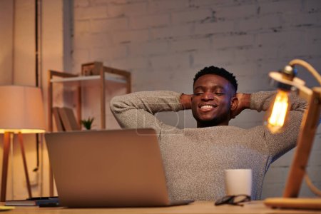 Téléchargez les photos : Heureux et détendu homme afro-américain regardant ordinateur portable dans le bureau à la maison la nuit, pigiste - en image libre de droit