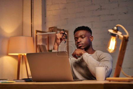 Téléchargez les photos : Réfléchi afro-américain pigiste tenant des lunettes de vue regardant ordinateur portable dans le bureau à la maison la nuit - en image libre de droit