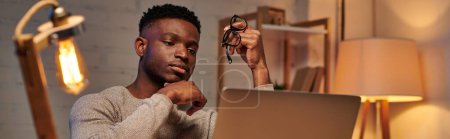 Photo for Pensive african american freelancer holding eyeglasses looking at laptop in home office, banner - Royalty Free Image