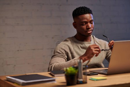 Téléchargez les photos : Fatigué afro-américain freelance tenant des lunettes tout en étant assis à l'ordinateur portable dans le bureau à la maison la nuit - en image libre de droit