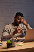exhausted african american freelancer sitting with eyeglasses near laptop in home office at night magic mug #692608388
