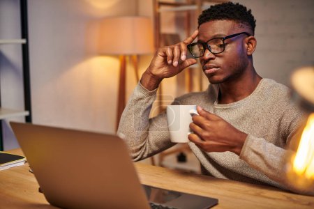 Téléchargez les photos : Travailleur indépendant afro-américain réfléchi avec tasse de café assis près d'un ordinateur portable la nuit au bureau à la maison - en image libre de droit