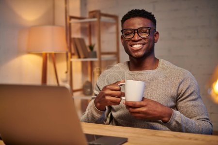 joyeux freelance afro-américain avec tasse de café assis près d'un ordinateur portable la nuit au bureau à la maison