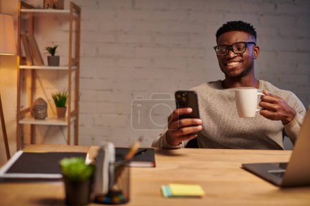 heureux freelance afro-américain avec tasse de café et smartphone près d'un ordinateur portable la nuit au bureau à domicile