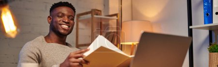 Téléchargez les photos : Heureux afro-américain pigiste avec ordinateur portable regardant dans le bureau à la maison la nuit, bannière - en image libre de droit