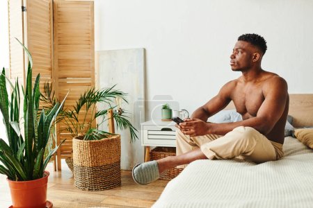 Foto de Hombre afroamericano reflexivo con teléfono inteligente sentado y mirando hacia otro lado en el dormitorio con decoración verde - Imagen libre de derechos