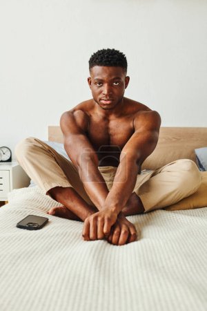 confident and strong african american man in pajama pants sitting on bed and looking at camera