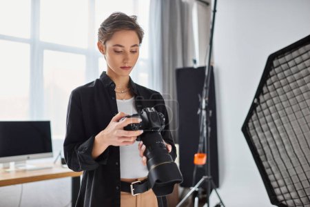 Foto de Joven atractiva fotógrafa en traje casual mirando fotos en su cámara en el estudio - Imagen libre de derechos