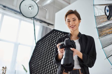 freudige junge Frau mit kurzen Haaren in Alltagskleidung posiert in ihrem Atelier mit der Kamera in der Hand