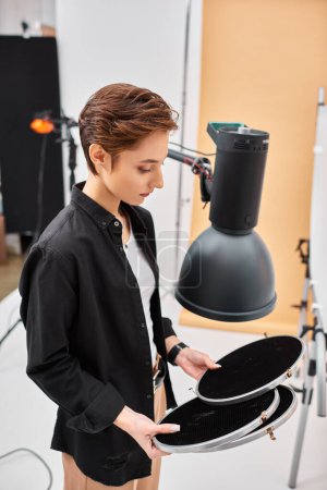 junge hübsche brünette Frau mit kurzen Haaren betrachtet ihre Fotoausrüstung in ihrem Atelier