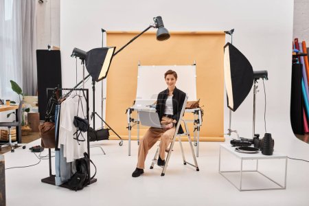 Téléchargez les photos : Joyeux jolie femme assise avec ordinateur portable tout en retouchant les photos et souriant à la caméra en studio - en image libre de droit