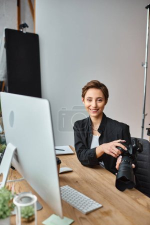 joyous attractive female photographer holding camera while retouching photos and smiling at camera