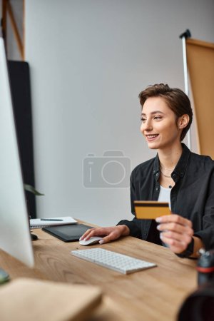 Téléchargez les photos : Heureux attrayant photographe femme payant en ligne en utilisant sa carte de crédit et souriant joyeusement - en image libre de droit
