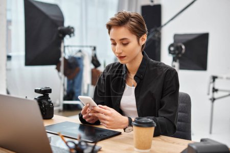 atractiva joven mujer de pelo corto con teléfono inteligente en sus manos mientras trabaja en un estudio de fotografía