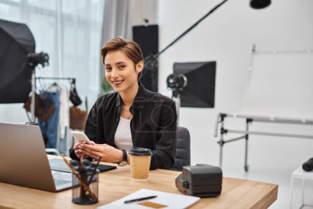 cheerful woman with smartphone in hands looking happily at camera while working in photo studio