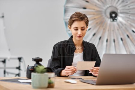 Photo for Attractive female photographer in casual attire sitting at table and looking at photos in her hands - Royalty Free Image