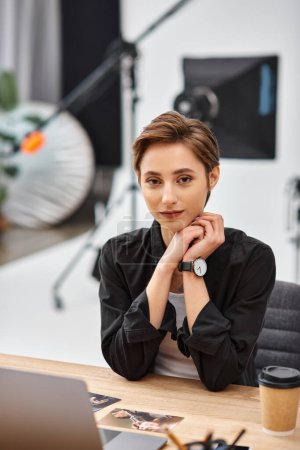 Photo for Young attractive woman in casual attire sitting at desk in her photo studio and looking at camera - Royalty Free Image