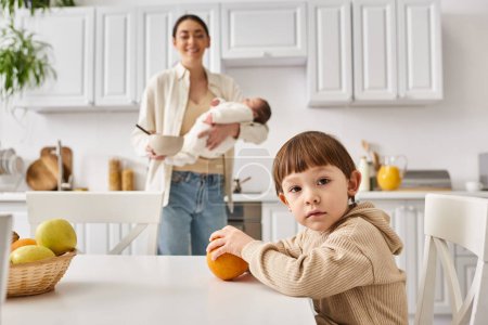 joyful mother in casual attire preparing breakfast for her toddler son while holding her newborn